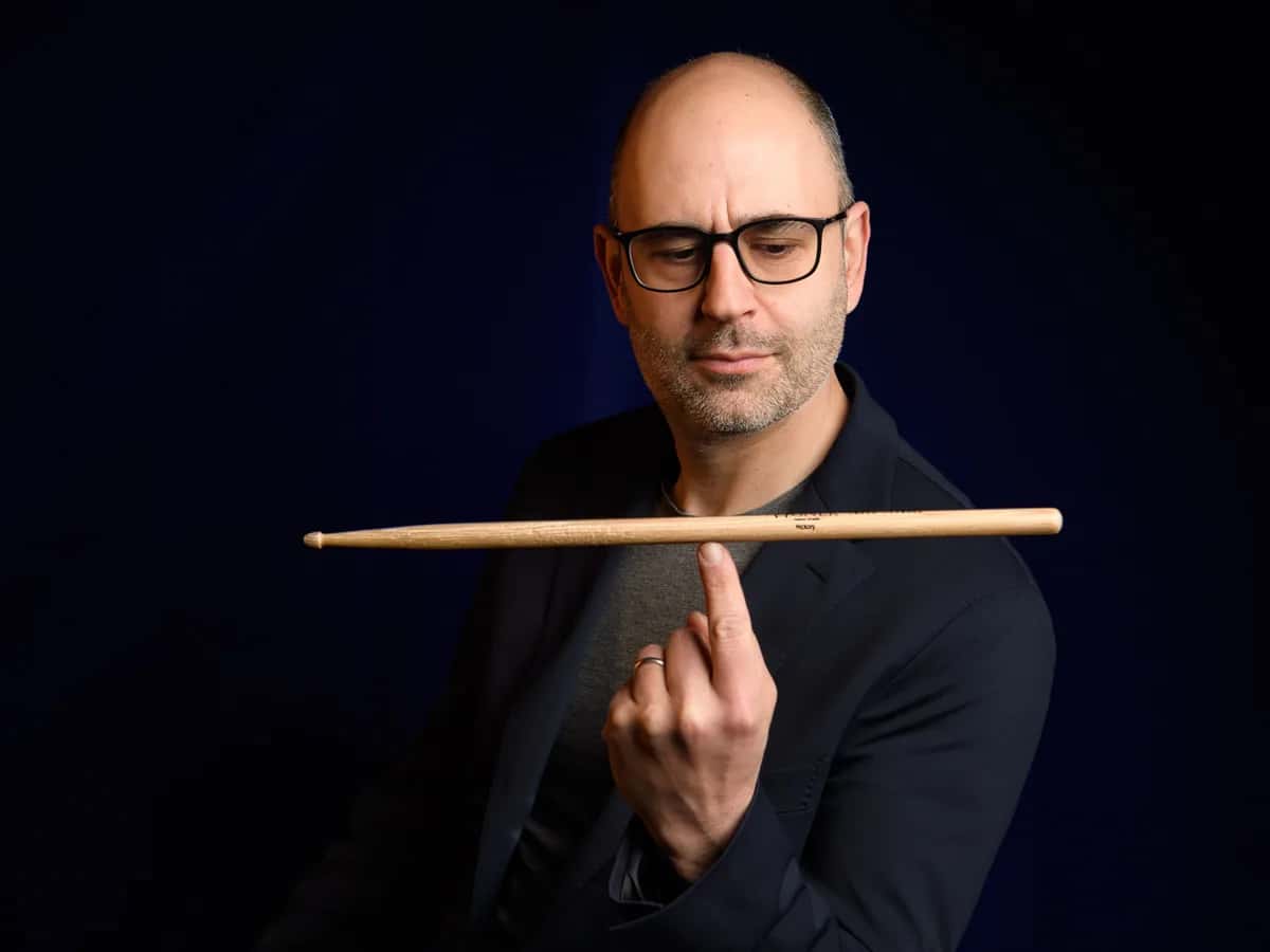 Paolo Orlandi, jazz drummer, balancing a drumstick on one finger against a dark blue background